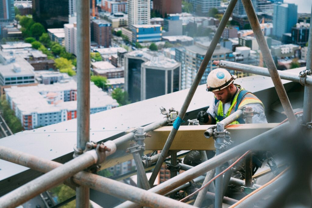Construction worker wearing a safety vest and helmet is working on scaffolding high above a cityscape.