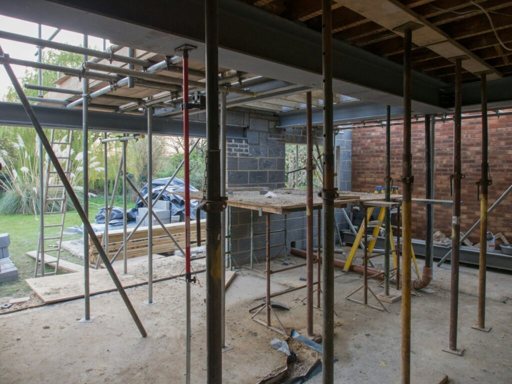 A room under construction features scaffolding poles, wooden planks, and tools, prioritizing construction site safety. Exposed brick walls and a partially open ceiling offer a glimpse of the garden beyond.