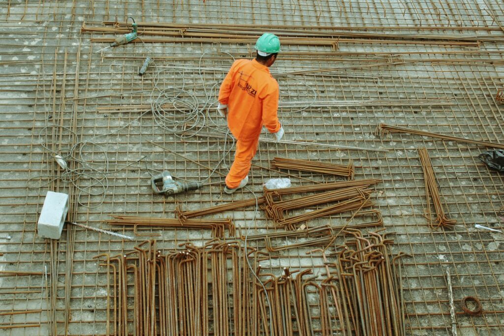 A construction worker in an orange uniform and green helmet navigates the rebar grid at a bustling site, surrounded by metal rods and cables, all under the watchful eye of general contractor safety protocols.