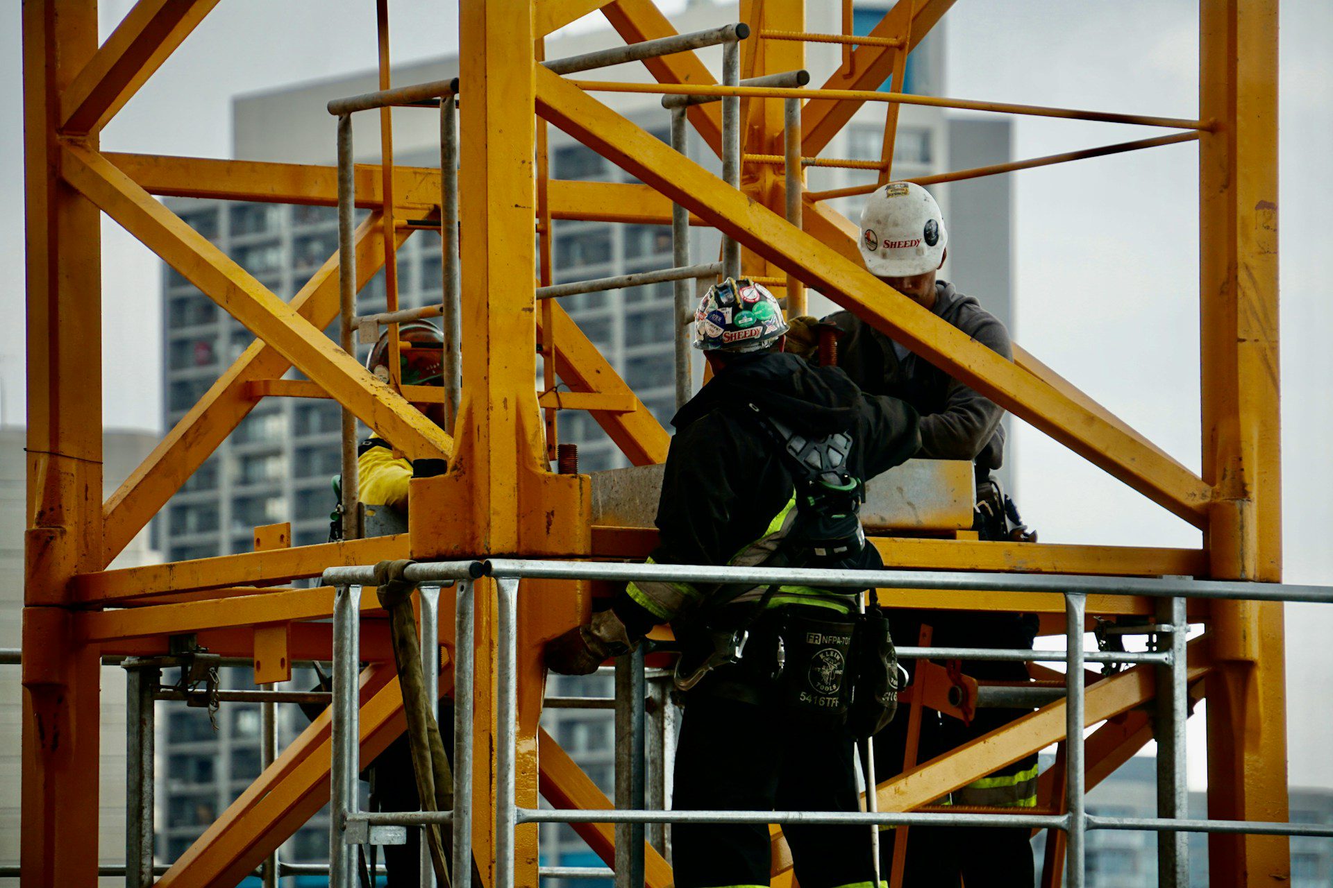 Three construction workers, assisted by general contractor safety software, are wearing safety gear as they assemble a yellow metal structure on a platform with city buildings in the background.