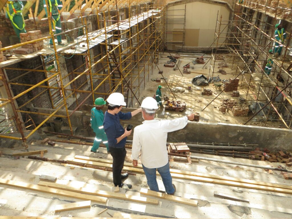 Two construction workers wearing hard hats stand on a site with scaffolding and building materials, discussing the progress. They're exploring ways to reduce admin in construction safety, aiming for streamlined processes without compromising on safety standards.