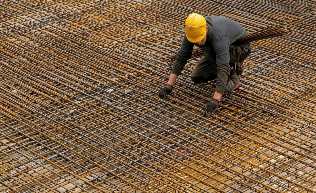 A worker, clad in a yellow helmet and gloves, skillfully maneuvers through a grid of steel rebar at a construction site. By adopting streamlined practices, the emphasis on reducing admin in construction safety becomes evident, ensuring efficiency without compromising security.