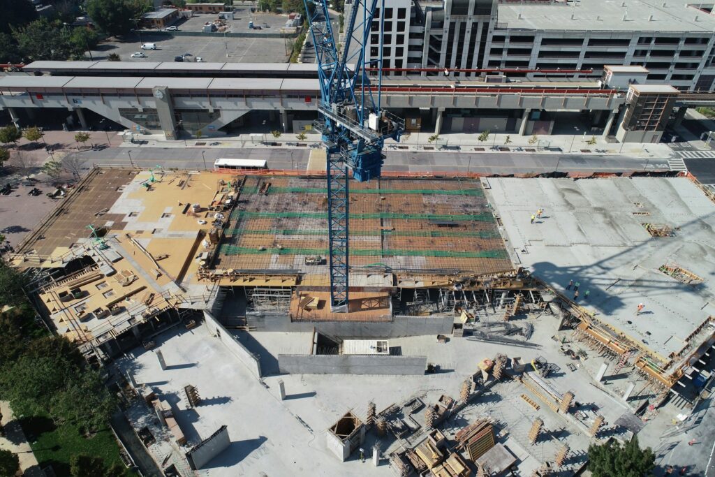 Aerial view of a construction site with a large blue crane at its heart, surrounded by partially built structures. Emphasizing construction site safety, the scene is framed by a parking garage in the background.