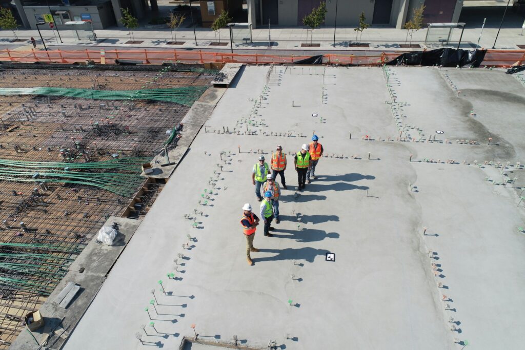 A group of construction workers in safety gear stand on a concrete slab surrounded by construction materials and unfinished areas.