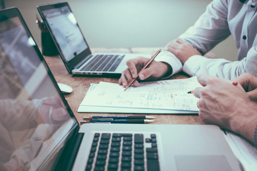 Two people collaborate at a desk with laptops and paperwork, aiming to reduce admin in construction safety. One person points with a pencil at a document, while the other listens attentively.